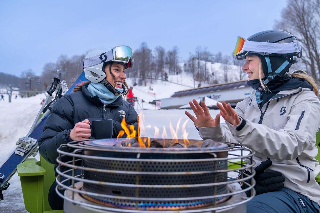Two skiers warm their hands over a metal fire pit in a snowy landscape.