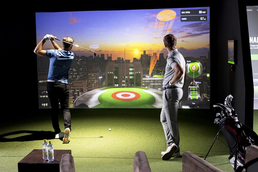 Two golfers practice swings before a virtual golf simulator screen.