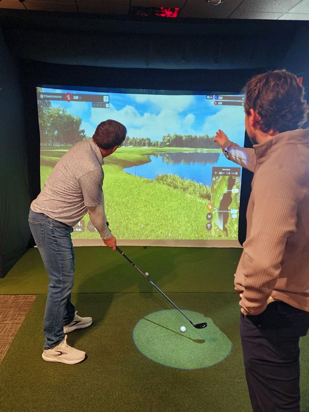 Two men playing indoor golf simulator, watching a virtual course on screen.